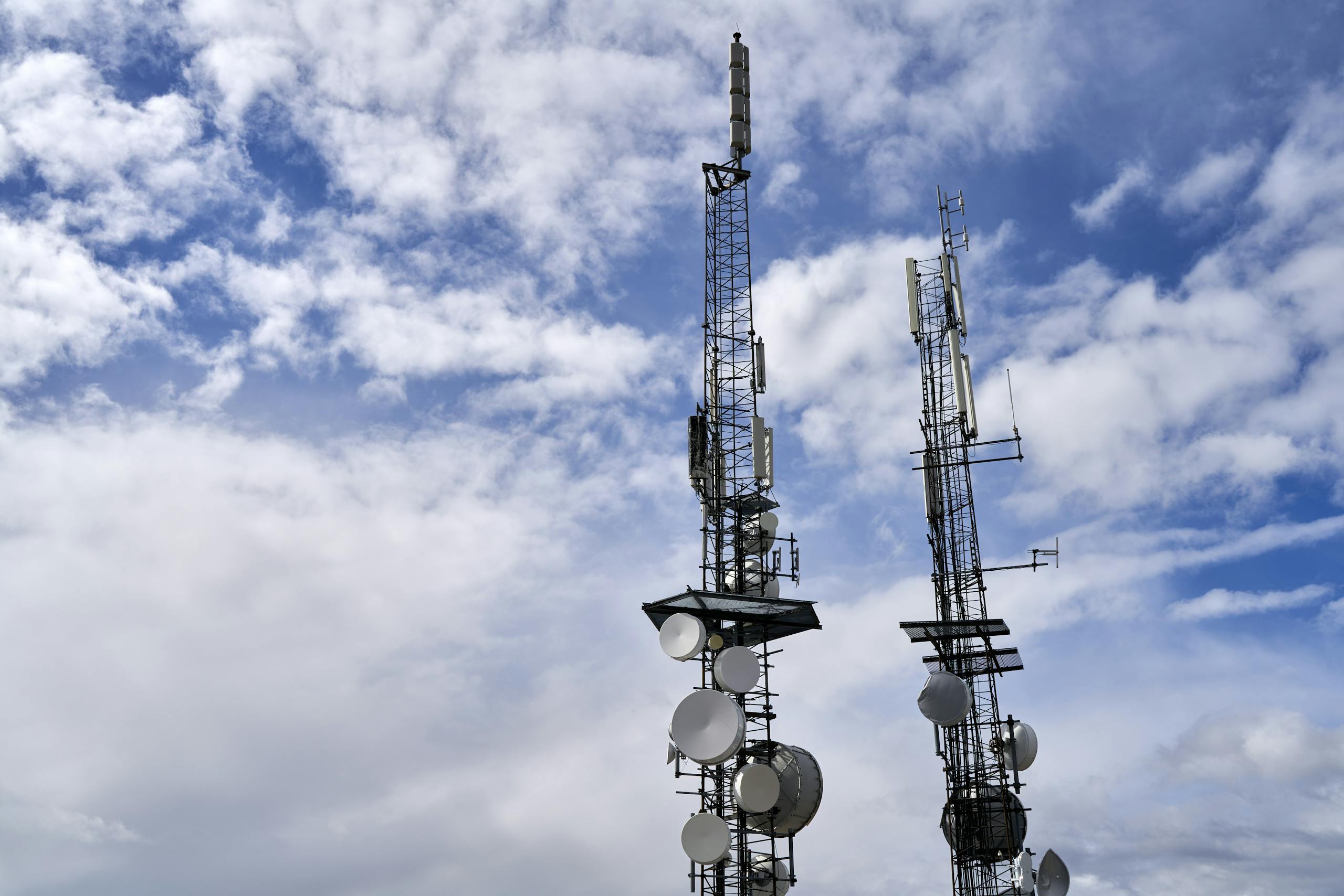 Telecommunication towers with antennas and dishes under a bright cloudy sky.